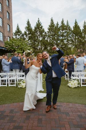 Joyful newlyweds celebrate after outdoor ceremony, bride in white gown and groom in navy suit pump fists in air.
