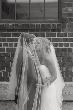 Black and white romantic photo of newlyweds kissing beneath flowing bridal veil outdoors.