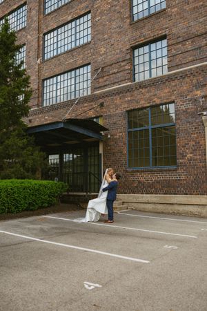 Bride and groom sharing romantic embrace in parking lot outside rustic brick industrial wedding venue.