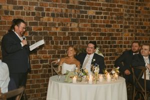 Wedding guest delivers a heartfelt speech to smiling newlyweds at a candlelit reception table.