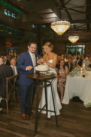 Bride and groom cutting wedding cake at elegant indoor reception with chandeliers and guests seated nearby.