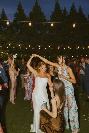 Bride in white gown dances with guests under warm string lights during outdoor evening wedding reception.