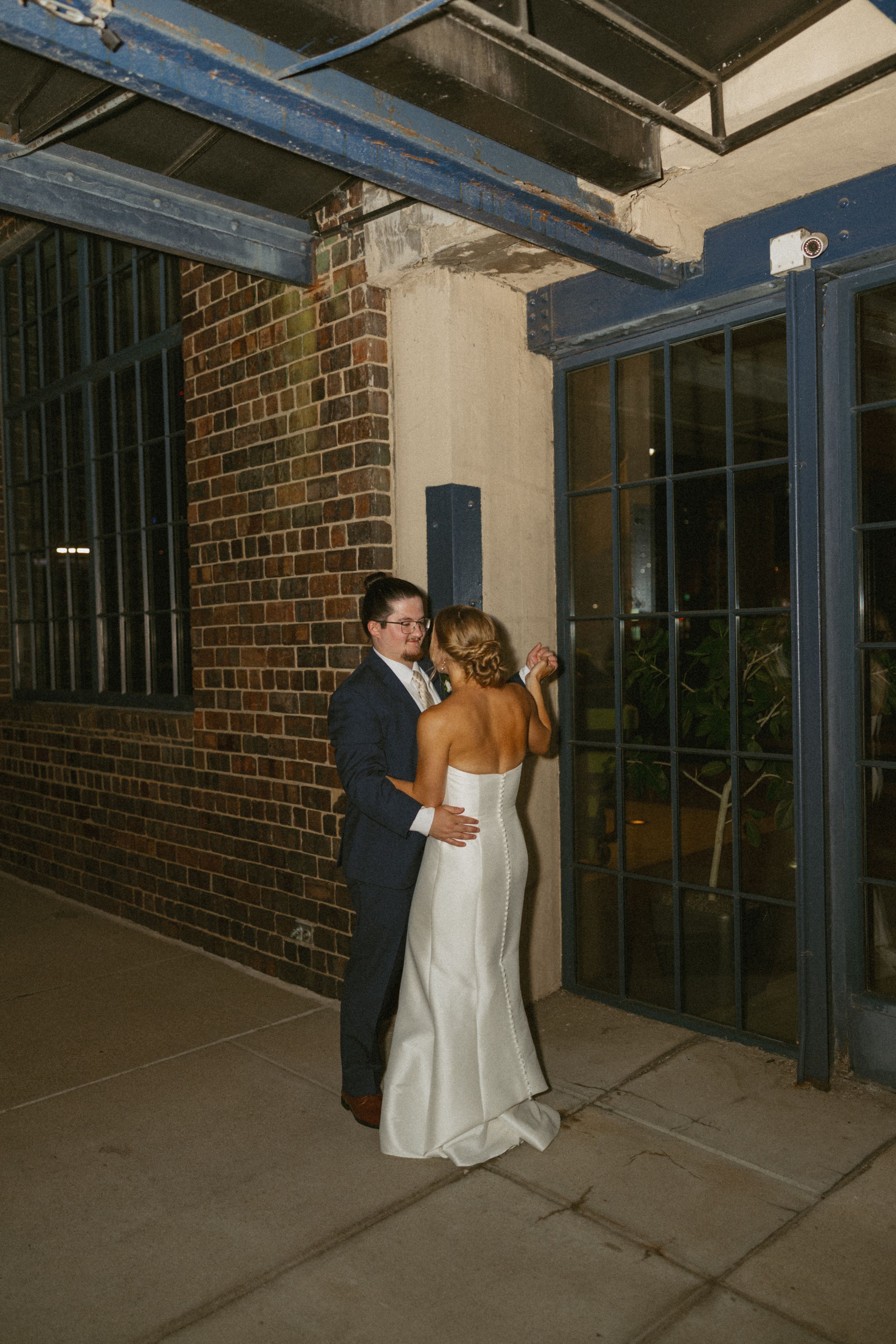 Bride and groom share intimate moment outside brick building with blue steel doors at night.
