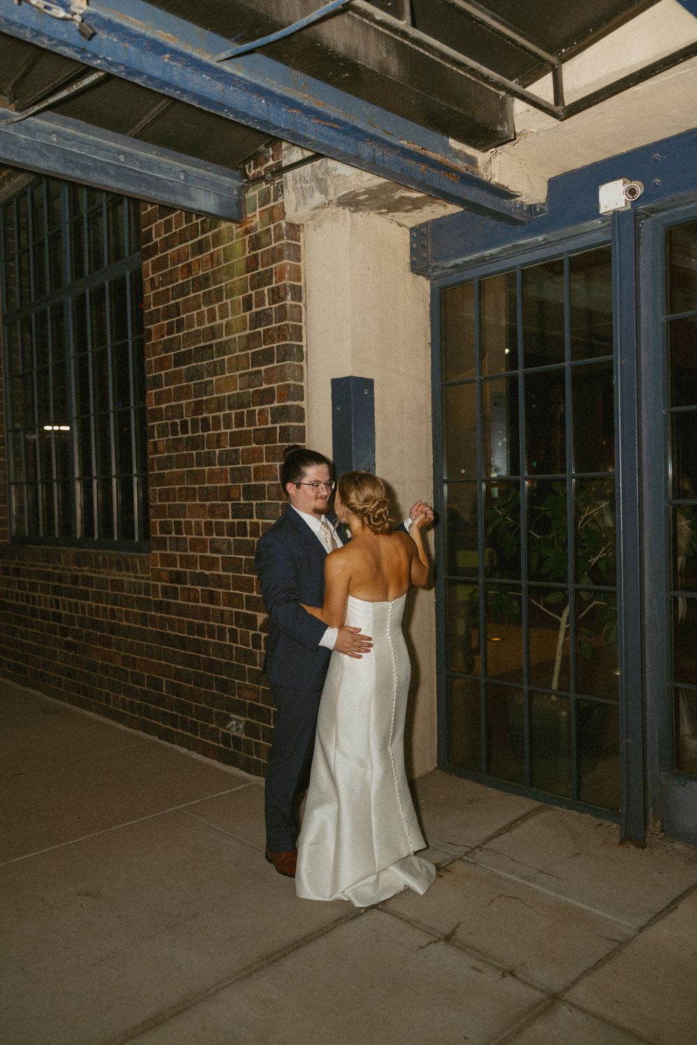 Bride and groom share intimate moment outside brick building with blue steel doors at night.