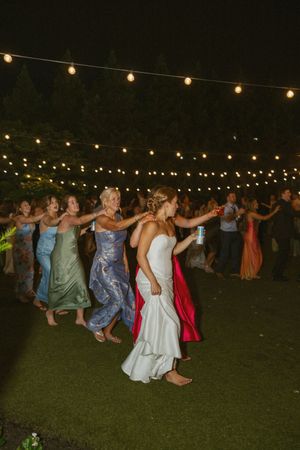 Bride leads a joyful conga line with guests outdoors under string lights at nighttime wedding reception.