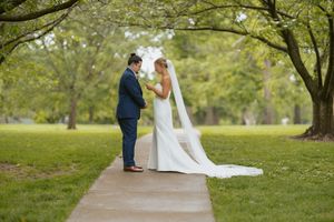 Bride and groom share a tender first look moment on a garden path surrounded by lush green trees.
