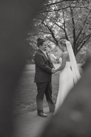 Black and white photo of bride and groom sharing first look moment on tree-lined path.