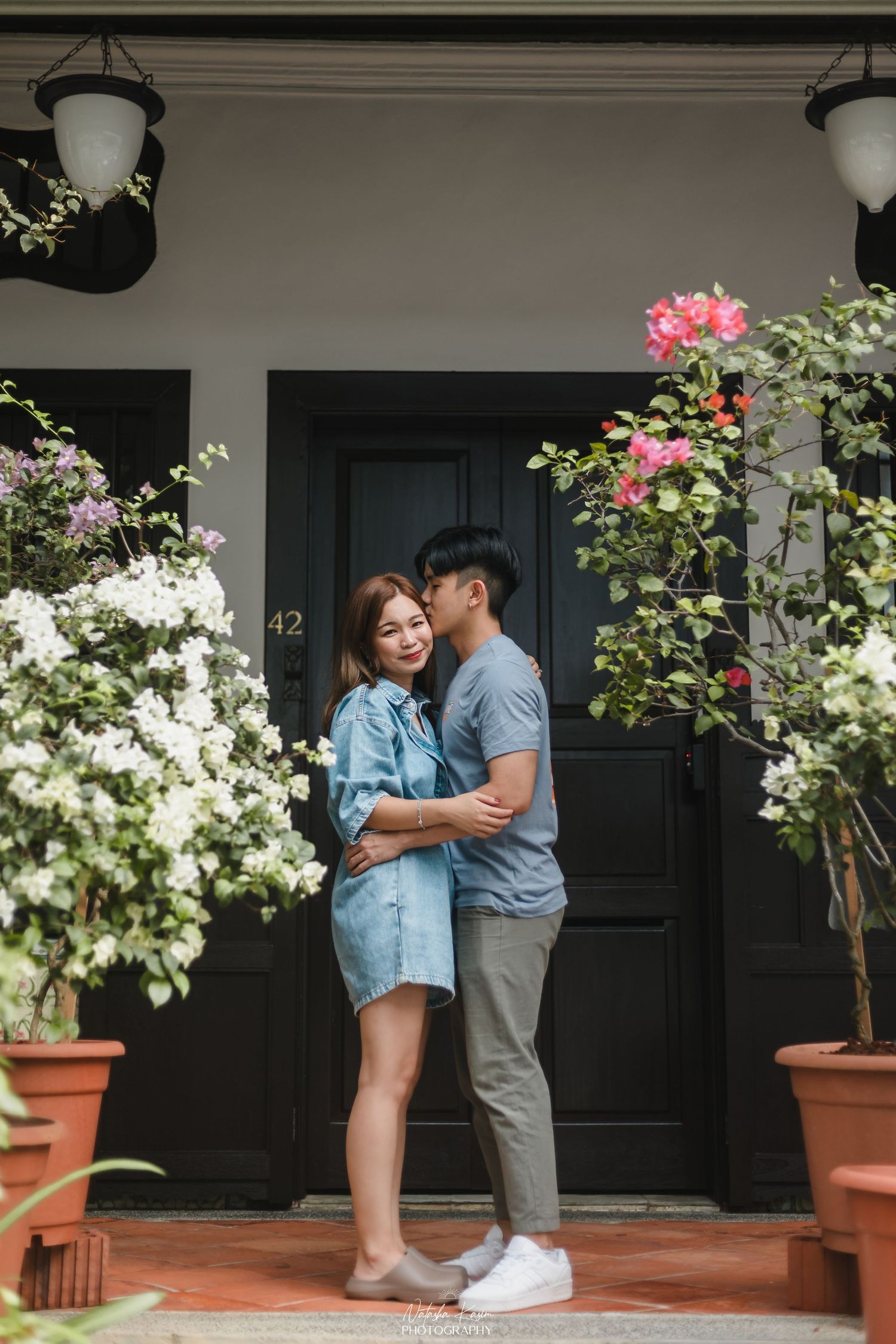 A couple embraces and shares a kiss in front of a black door surrounded by blooming white and pink flowers.