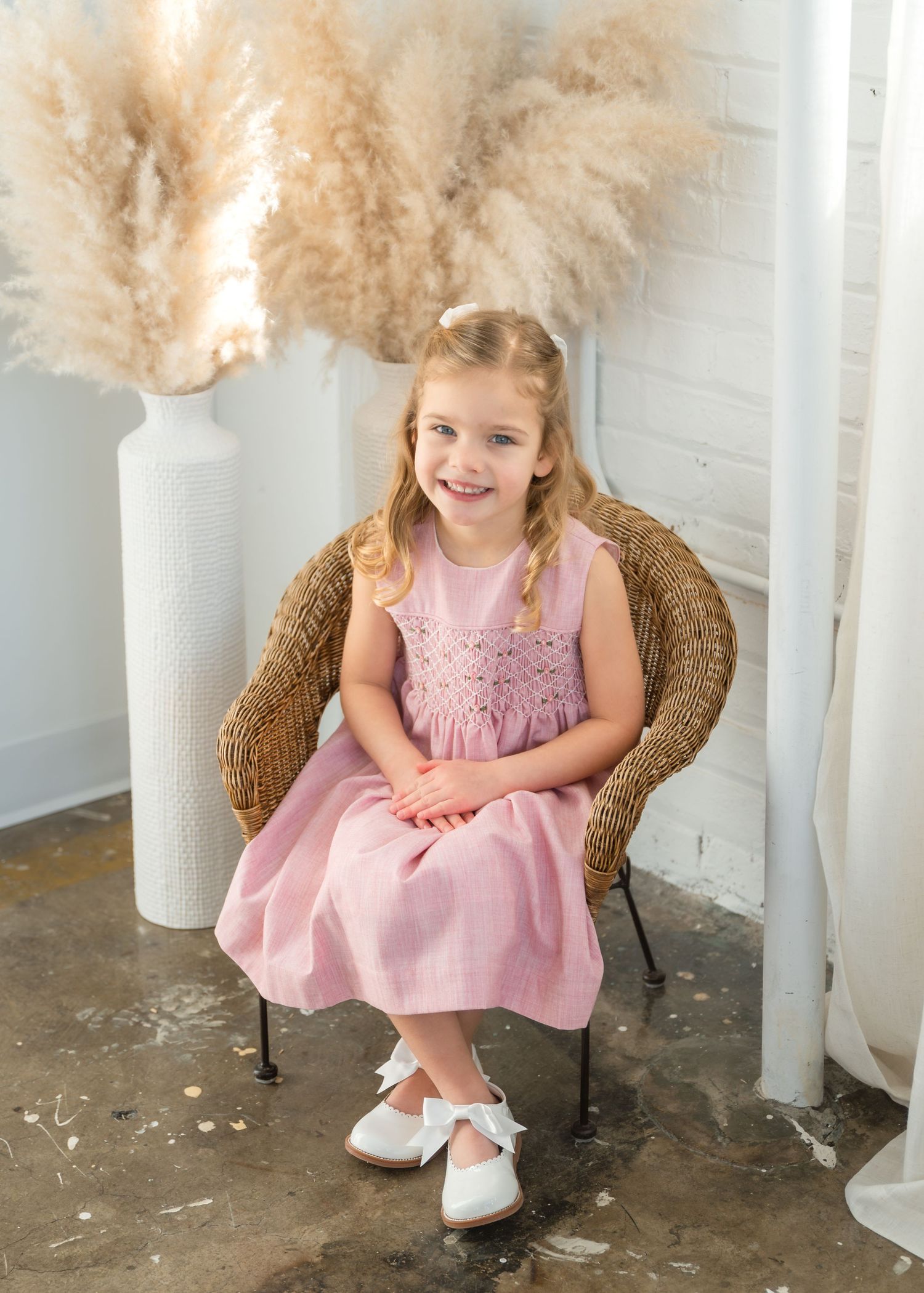 A smiling young girl in a pink smocked dress and white shoes sits in a wicker chair with pampas grass decor.