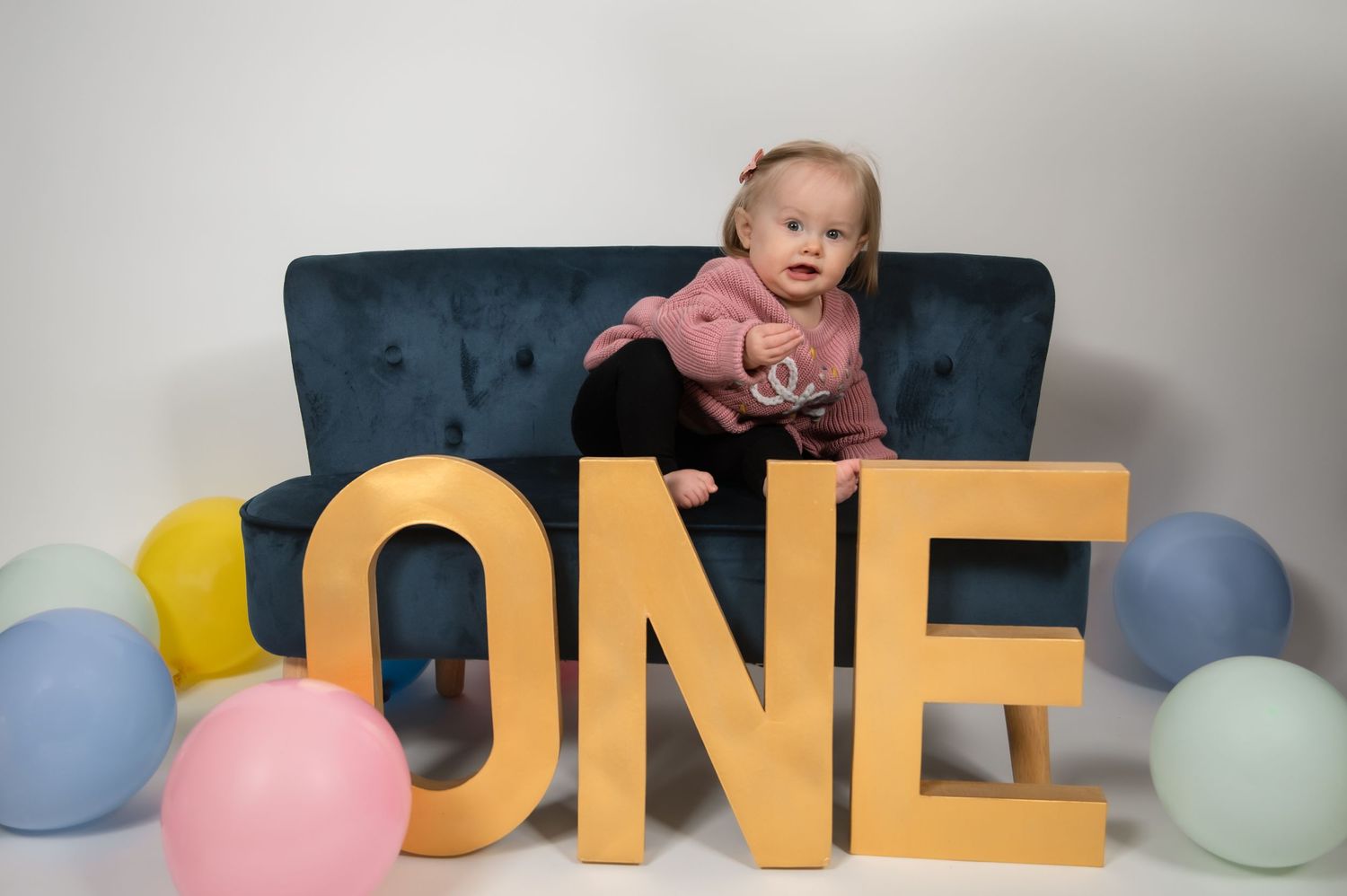 Baby girl celebrating her first birthday sitting on a blue sofa with large gold ONE letters and colorful balloons.