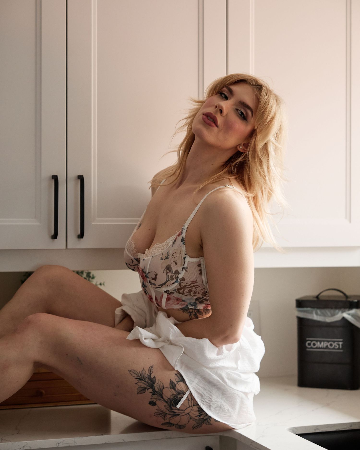 Blonde woman in floral camisole and white skirt sitting on kitchen counter with floral thigh tattoo.