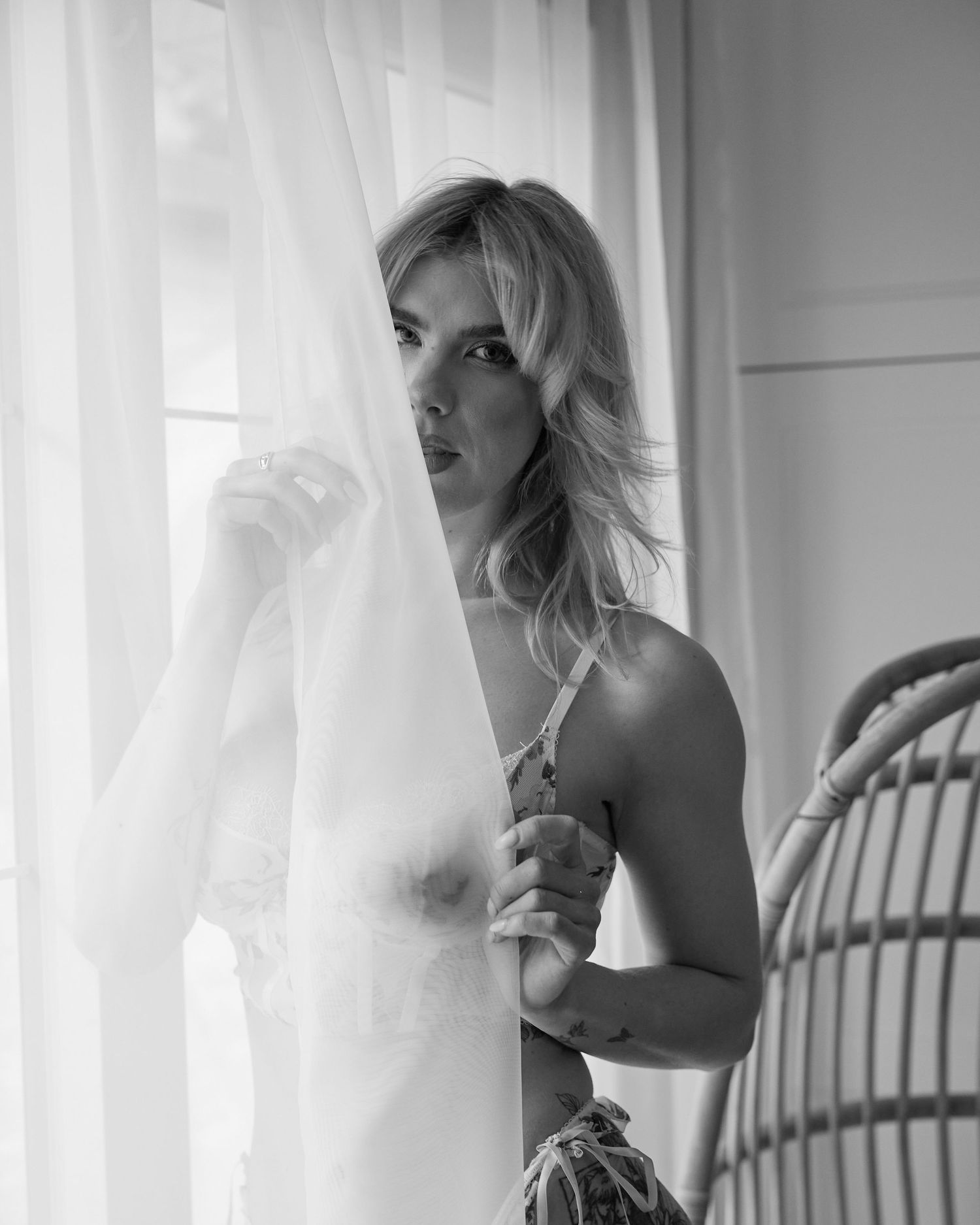 Black and white boudoir photo of a blonde woman peeking behind sheer white curtains near a bright window.