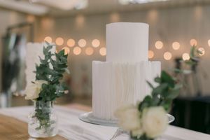 Elegant two-tier white wedding cake with textured ruffles, surrounded by greenery and roses on a table with bokeh lights.