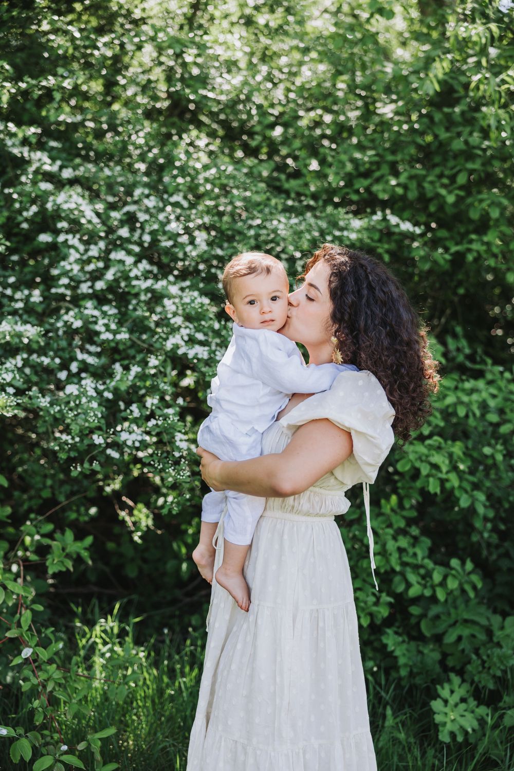 Mommy and me session - woman in a white dress kisses a baby boy outdoors among lush green flowering bushes in Nieuw Vennep.