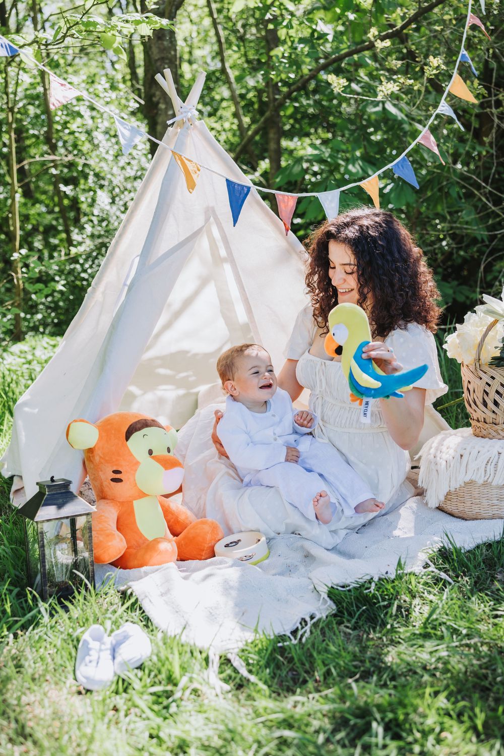 Mommy and me portrait of mom and baby playing outdoors near a teepee tent with colorful decoration, stuffed toys, and sunny green trees.