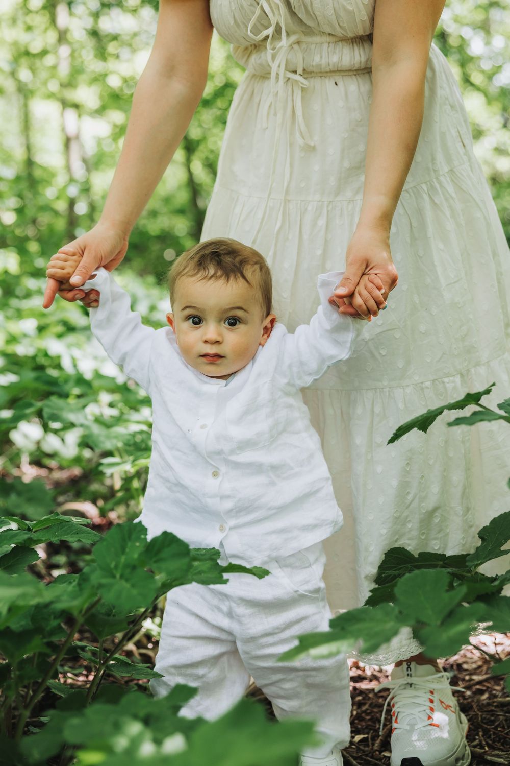 Portrait of baby in white outfit takes first steps outdoors, held by mother in white dress among lush green forest foliage.