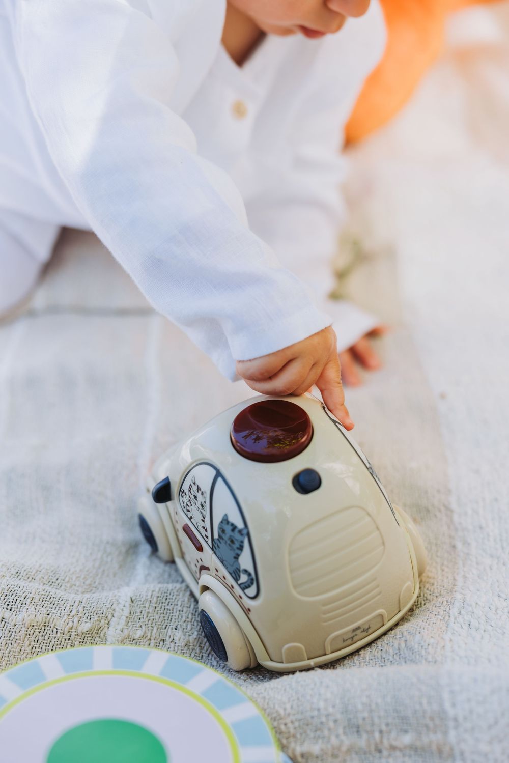 Young child in white outfit playing with a toy car on a blanket outdoors.