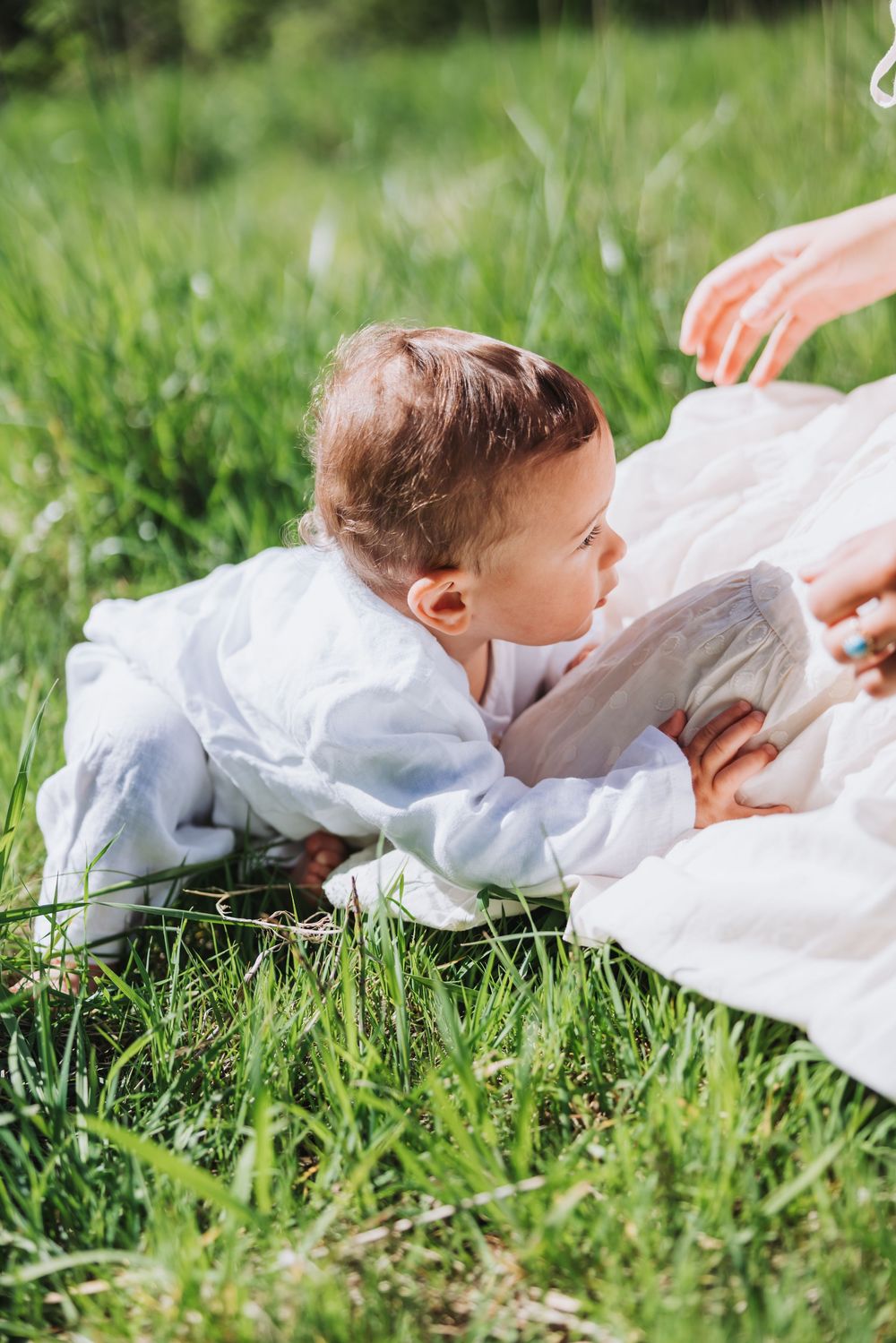 A baby in white clothing lies on a blanket in green grass outdoors on a sunny day.