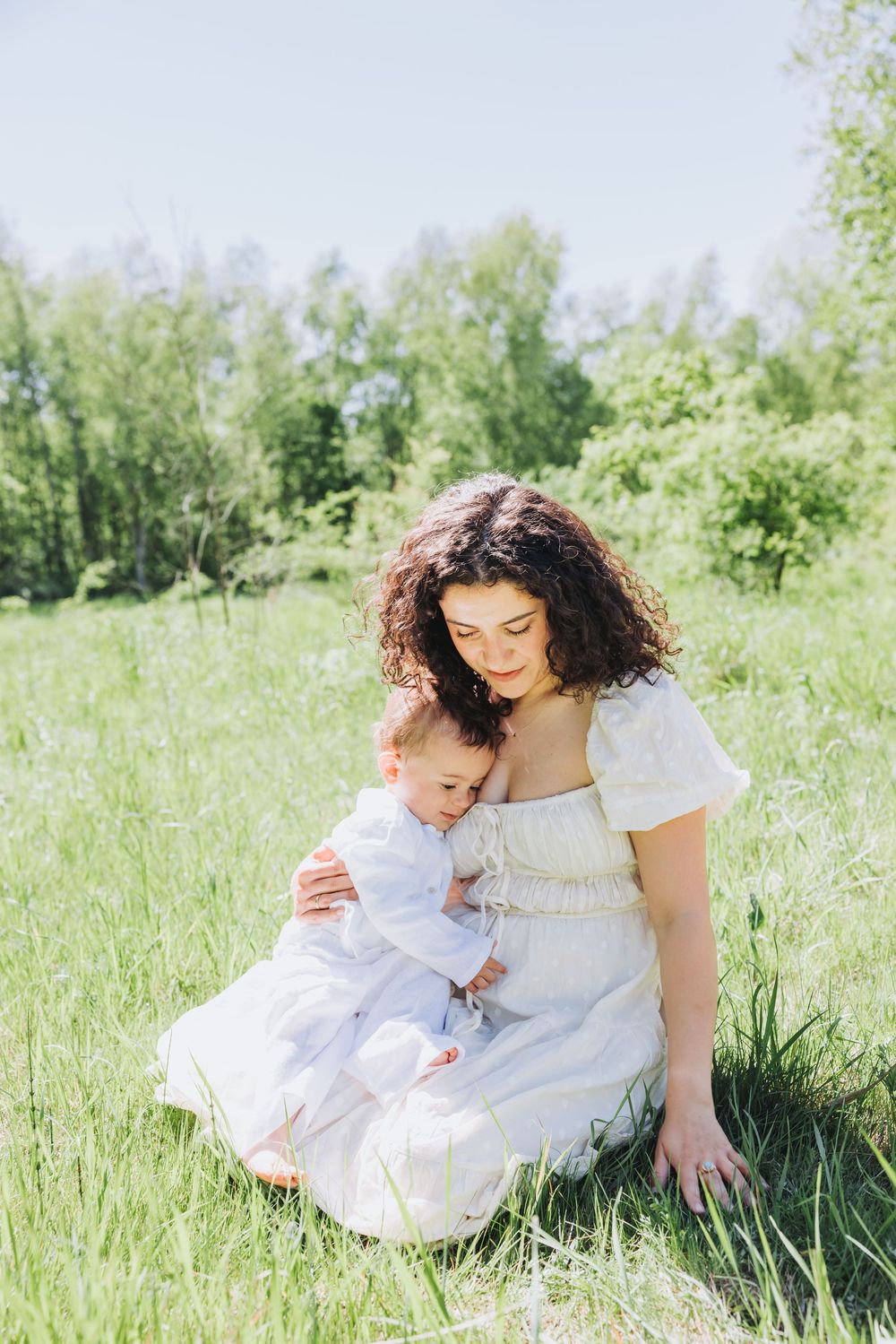 Candid photo of curly-haired mother in a white dress sitting on green grass, tenderly embracing her baby outdoors in Nieuw Vennep