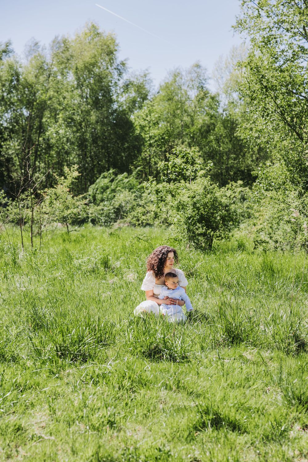 A curly-haired mother in white dress hugs her toddler on green grass meadow surrounded by lush trees on sunny day.
