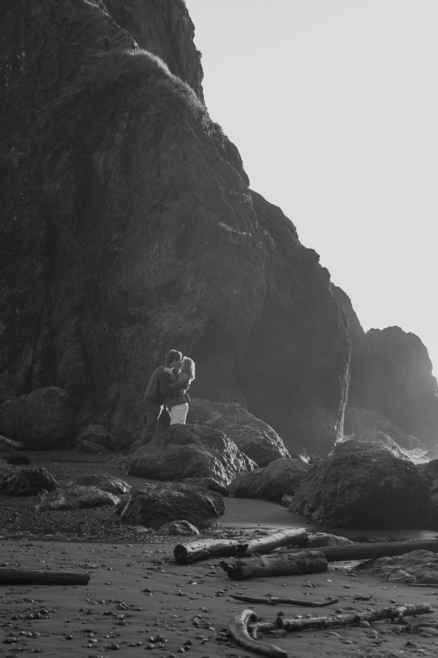 Black and white photo of a couple kissing on rocks at a misty beach with driftwood in the foreground.