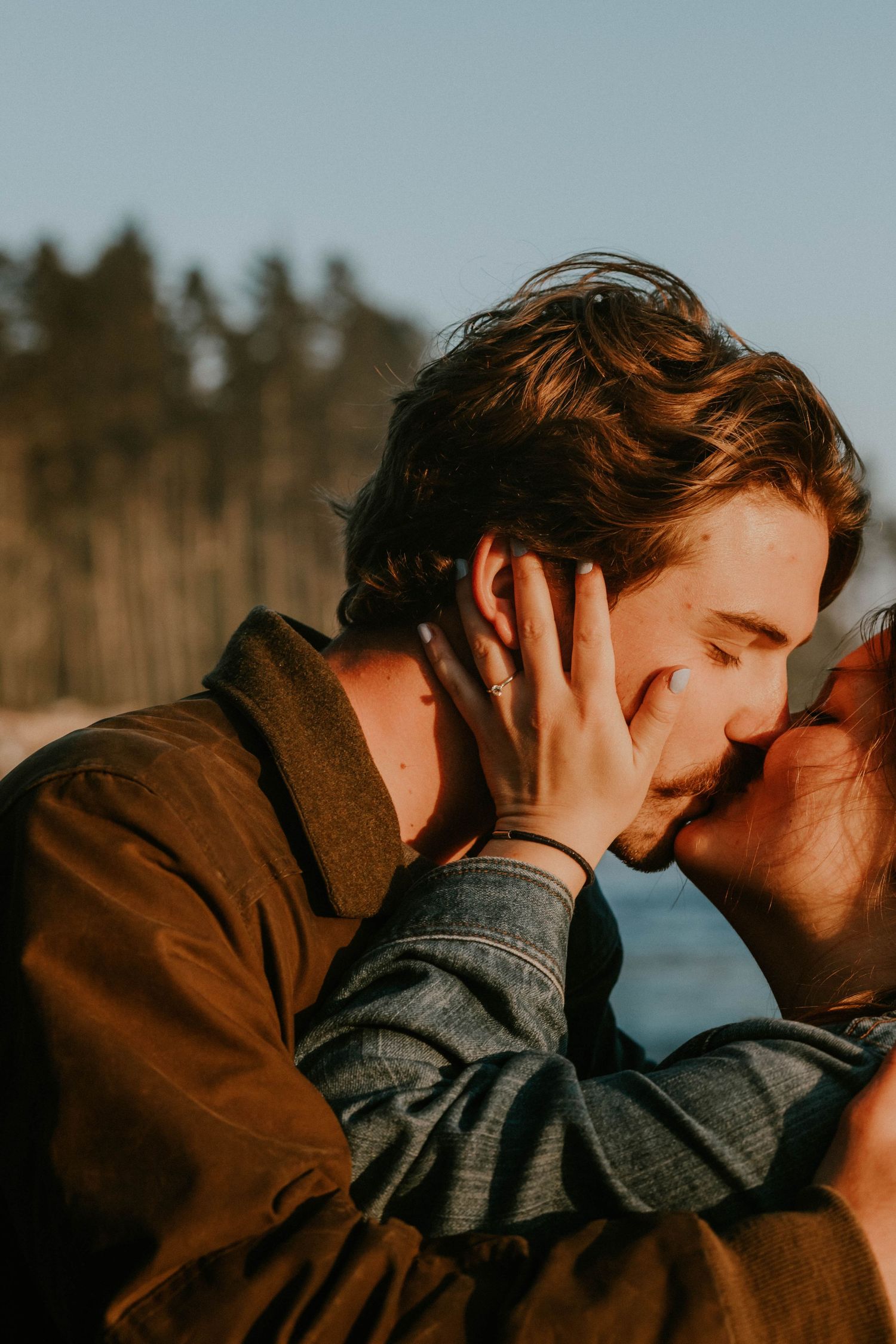 A couple shares a tender kiss outdoors at golden hour, with trees and blue sky in the background.