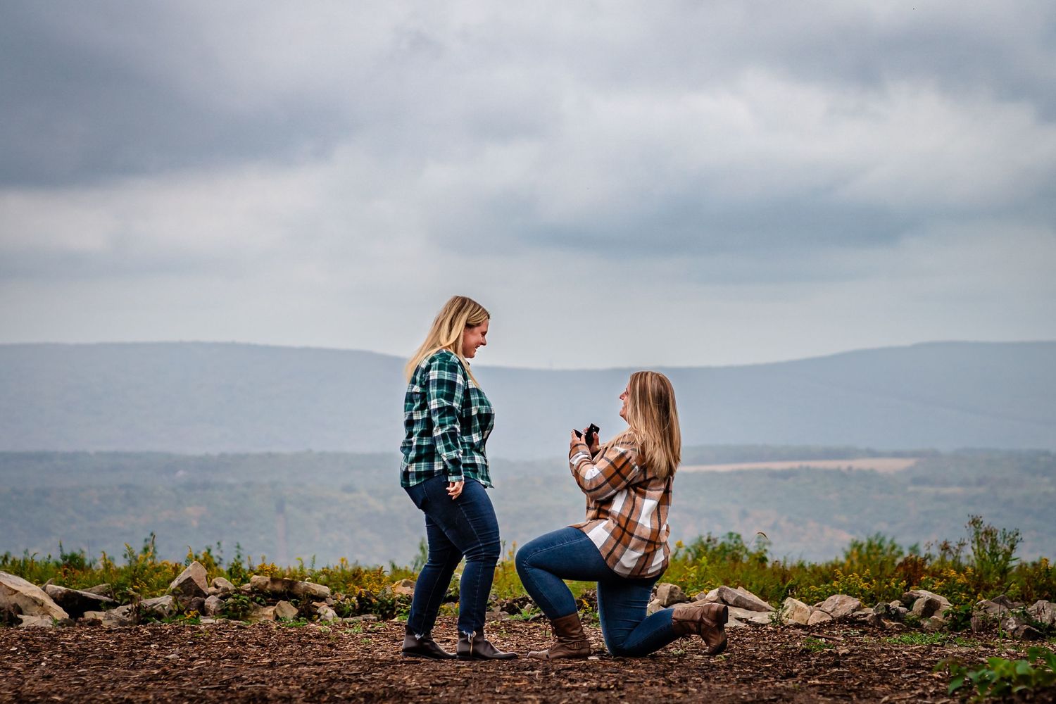 Two women in plaid shirts share a romantic proposal moment outdoors, with one kneeling holding a ring box.