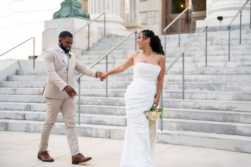 A Black bride and groom hold hands on grand steps, she in a white gown, he in a beige suit.