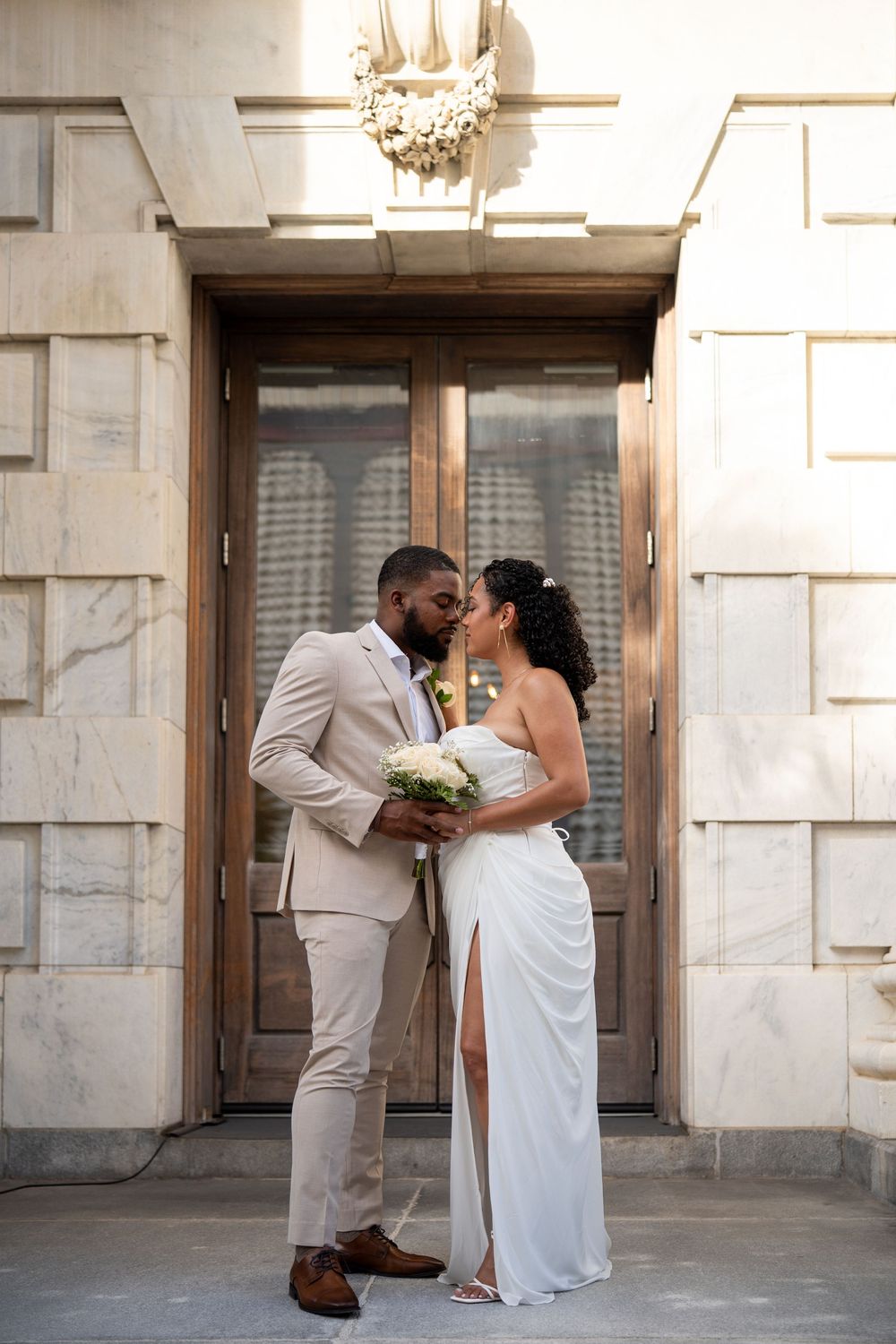 A bride and groom share a kiss outside an elegant stone building, she in a white gown, he in a tan suit holding her bouquet.