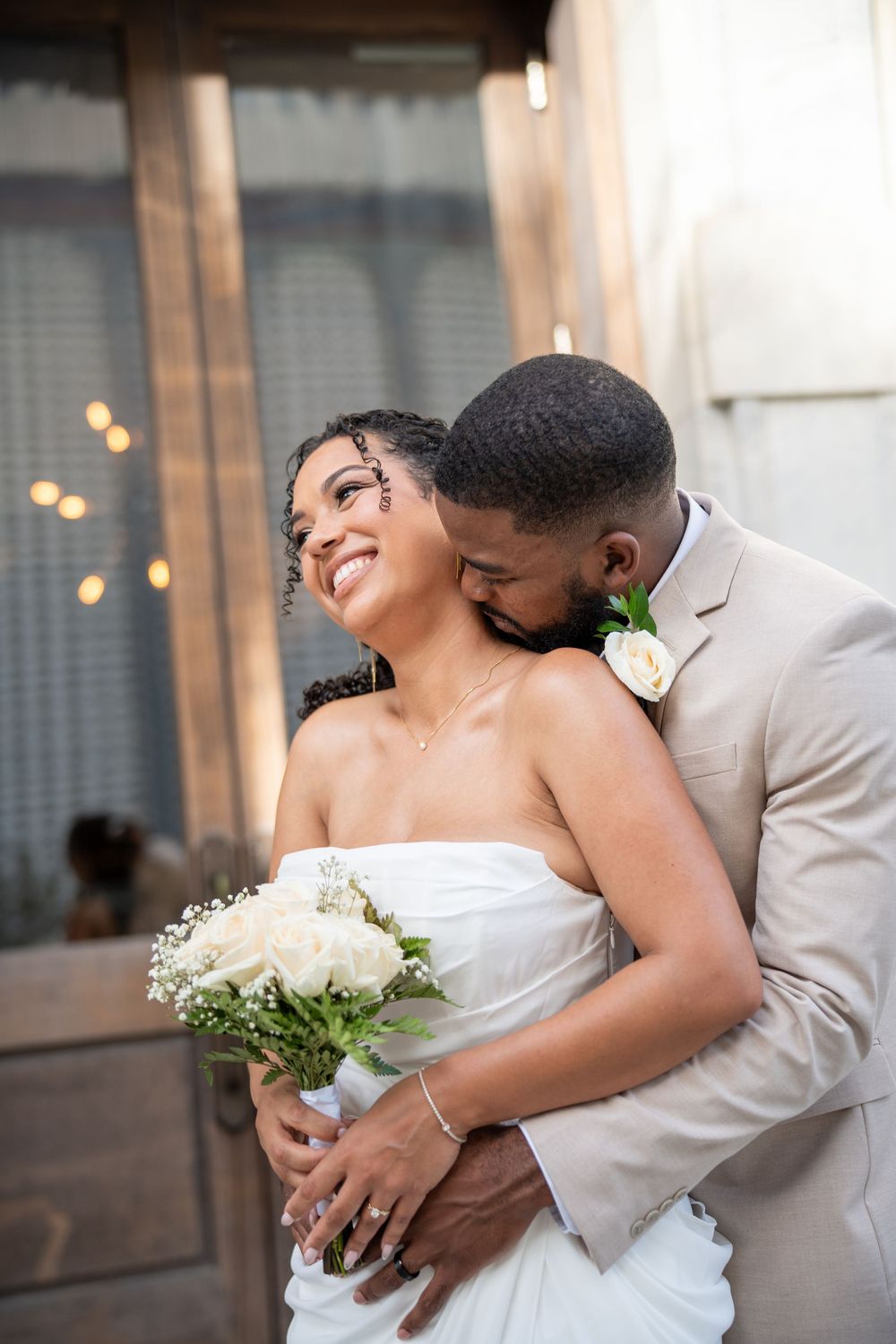 A joyful bride holds white roses as her groom in a tan suit embraces her from behind during their wedding day.