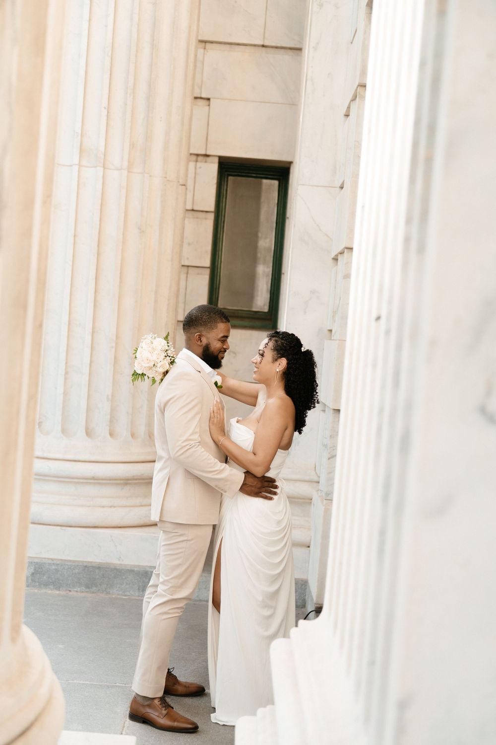 A bride and groom embrace between elegant marble columns, wearing white wedding attire with a floral bouquet.