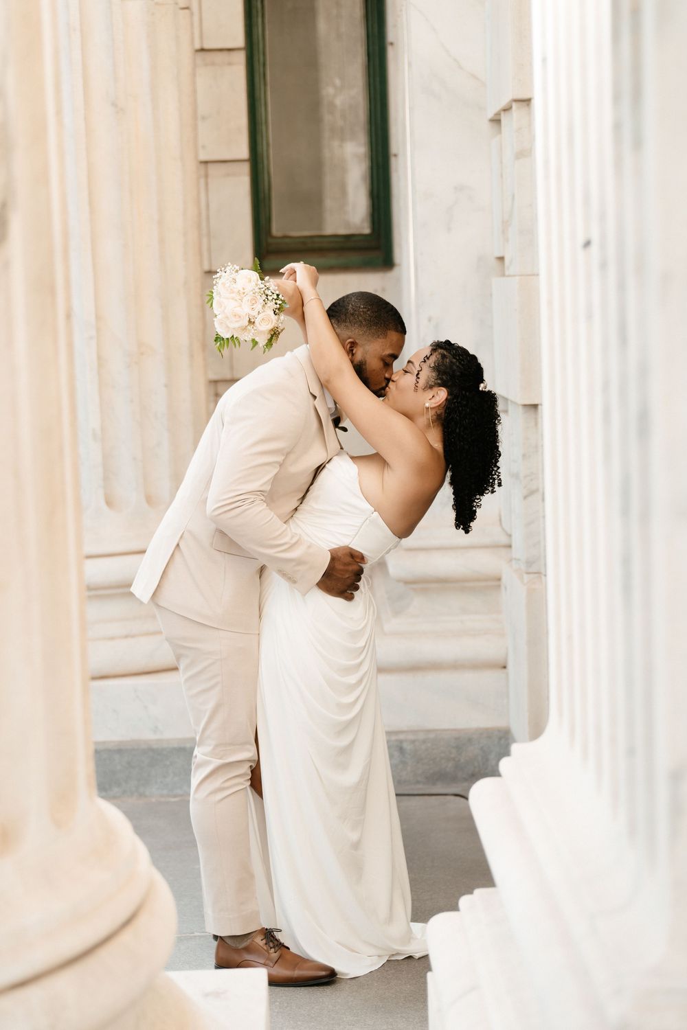 A bride and groom share a romantic dip kiss between white columns, she in a white gown, he in a cream suit.