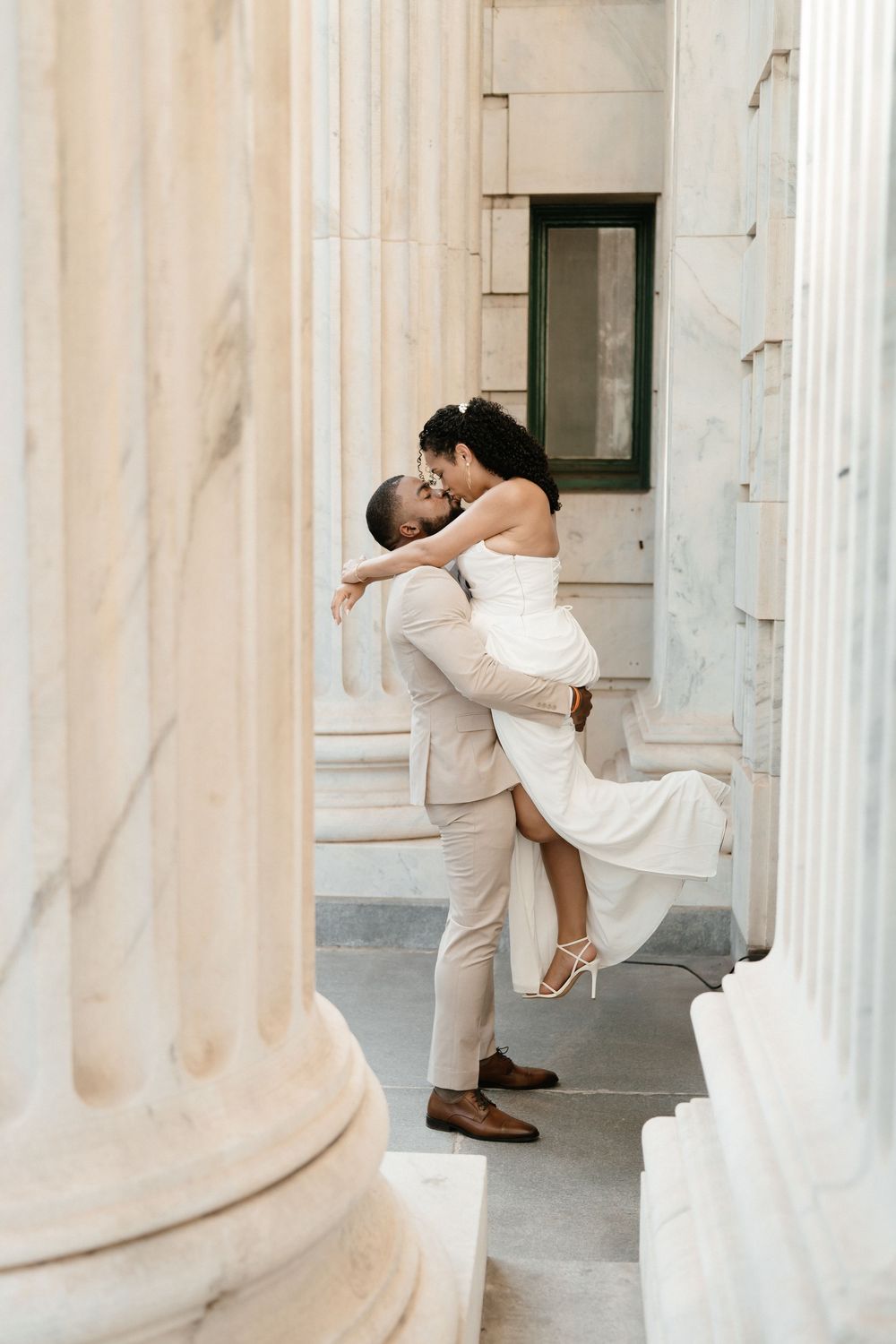 A groom lifts his bride in a white dress between marble columns in a romantic wedding photo.