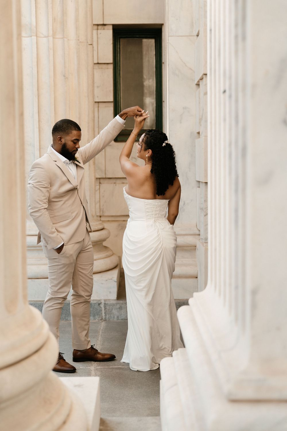 A groom in a beige suit twirls his bride in a white gown between elegant marble columns.