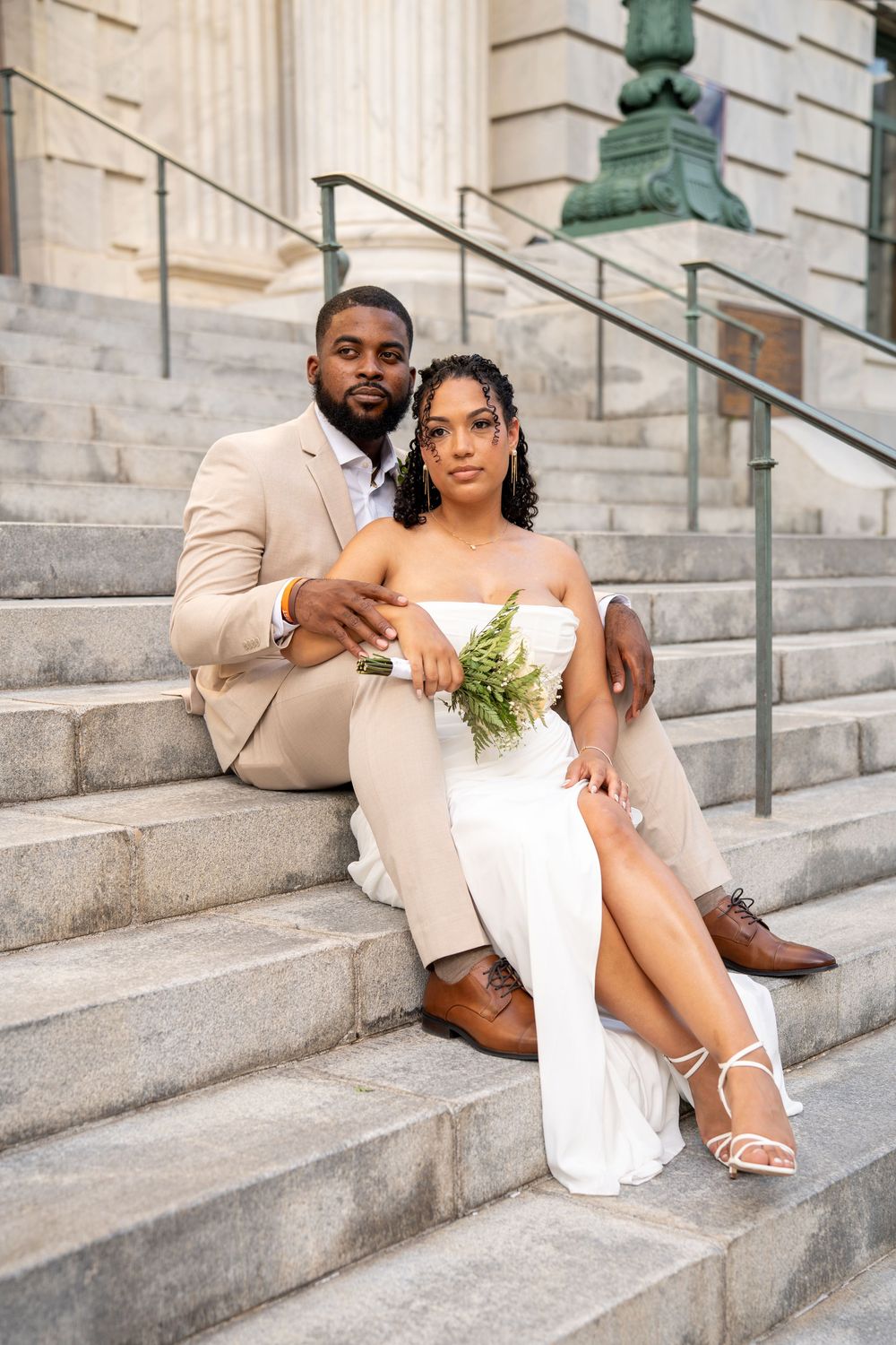 A couple in wedding attire sits on stone steps, the groom in a tan suit and bride in a white dress holding greenery.