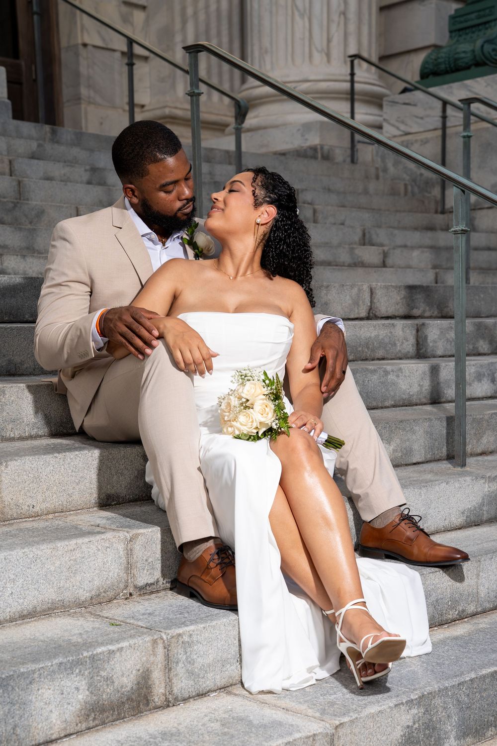 Bride and groom sitting on stone steps, sharing a romantic moment in wedding attire with bouquet.