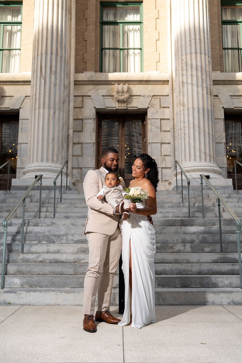 A newlywed couple in wedding attire holds their baby on the steps of an elegant stone building with tall columns.