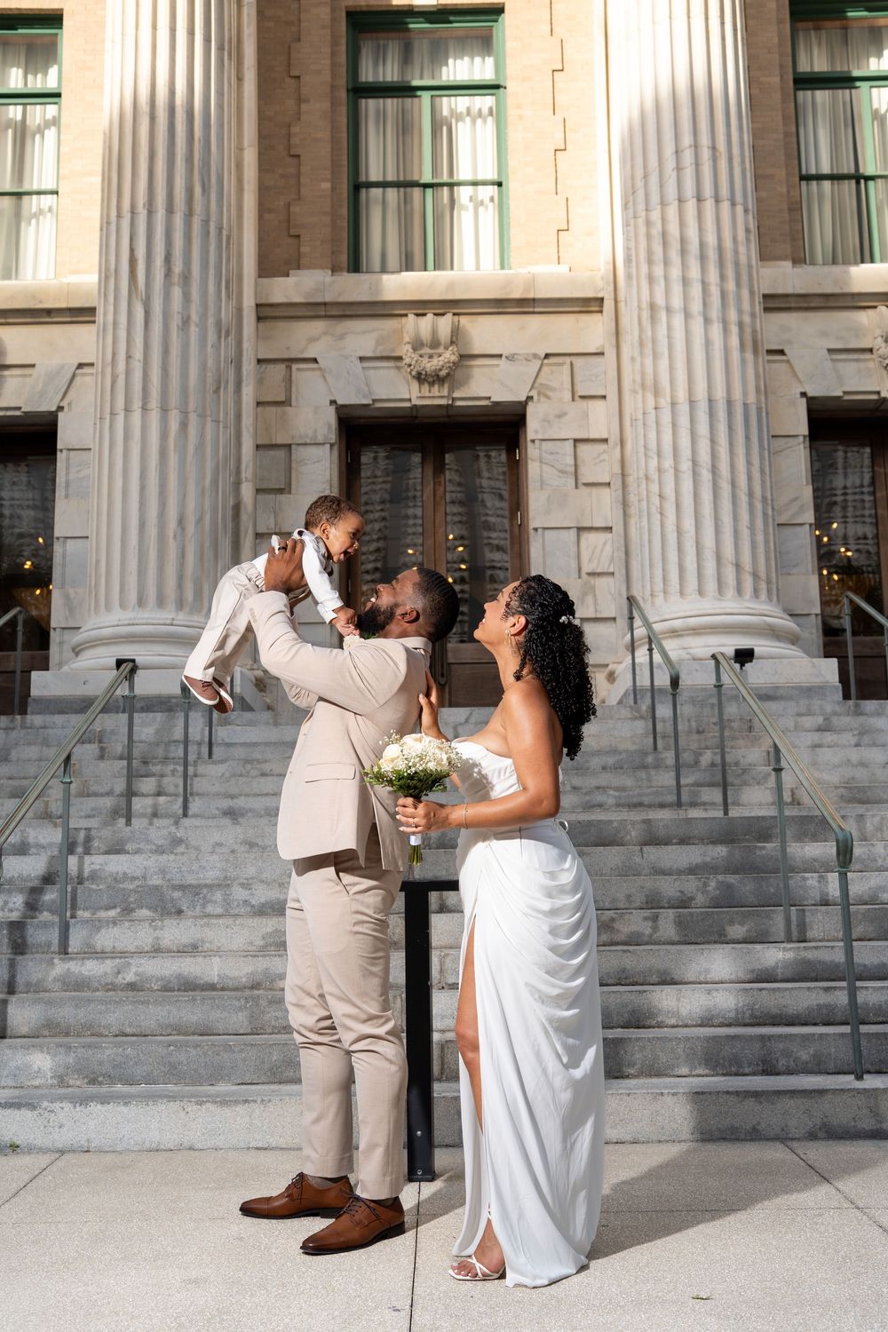 Newlywed couple in wedding attire lifting baby outside grand building with columns after courthouse wedding ceremony.