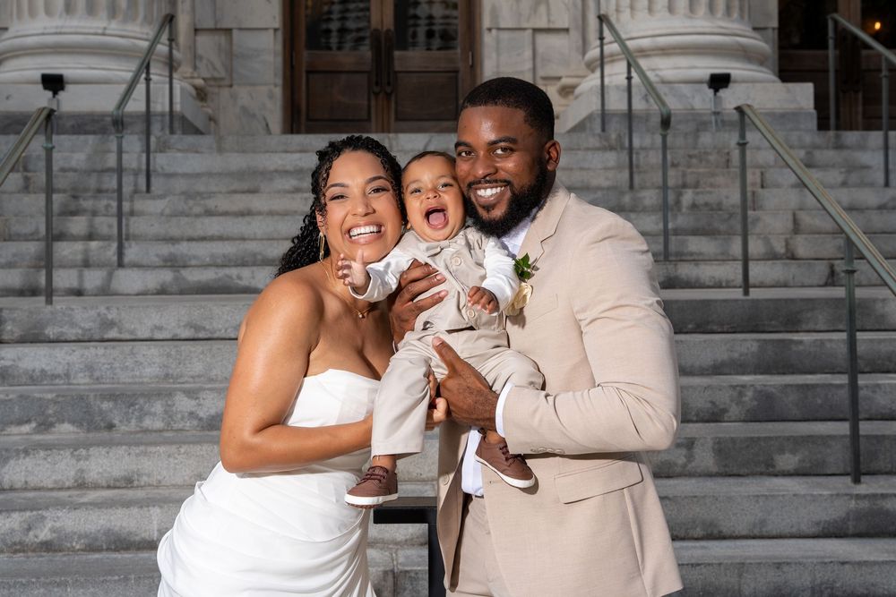 Happy couple in wedding attire holding their laughing baby on steps, bride in white gown, groom in beige suit.