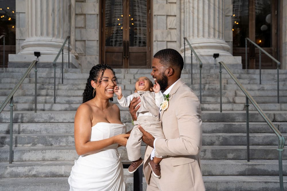 Newlywed couple in wedding attire holding baby on church steps, celebrating marriage and new family.
