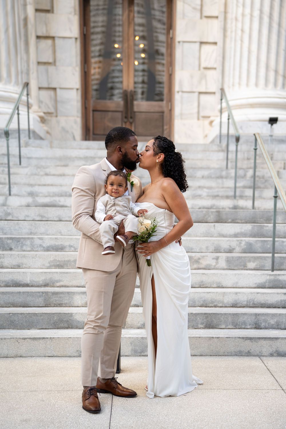 Newlywed couple kissing on courthouse steps, groom in beige suit holding baby, bride in white gown with bouquet.