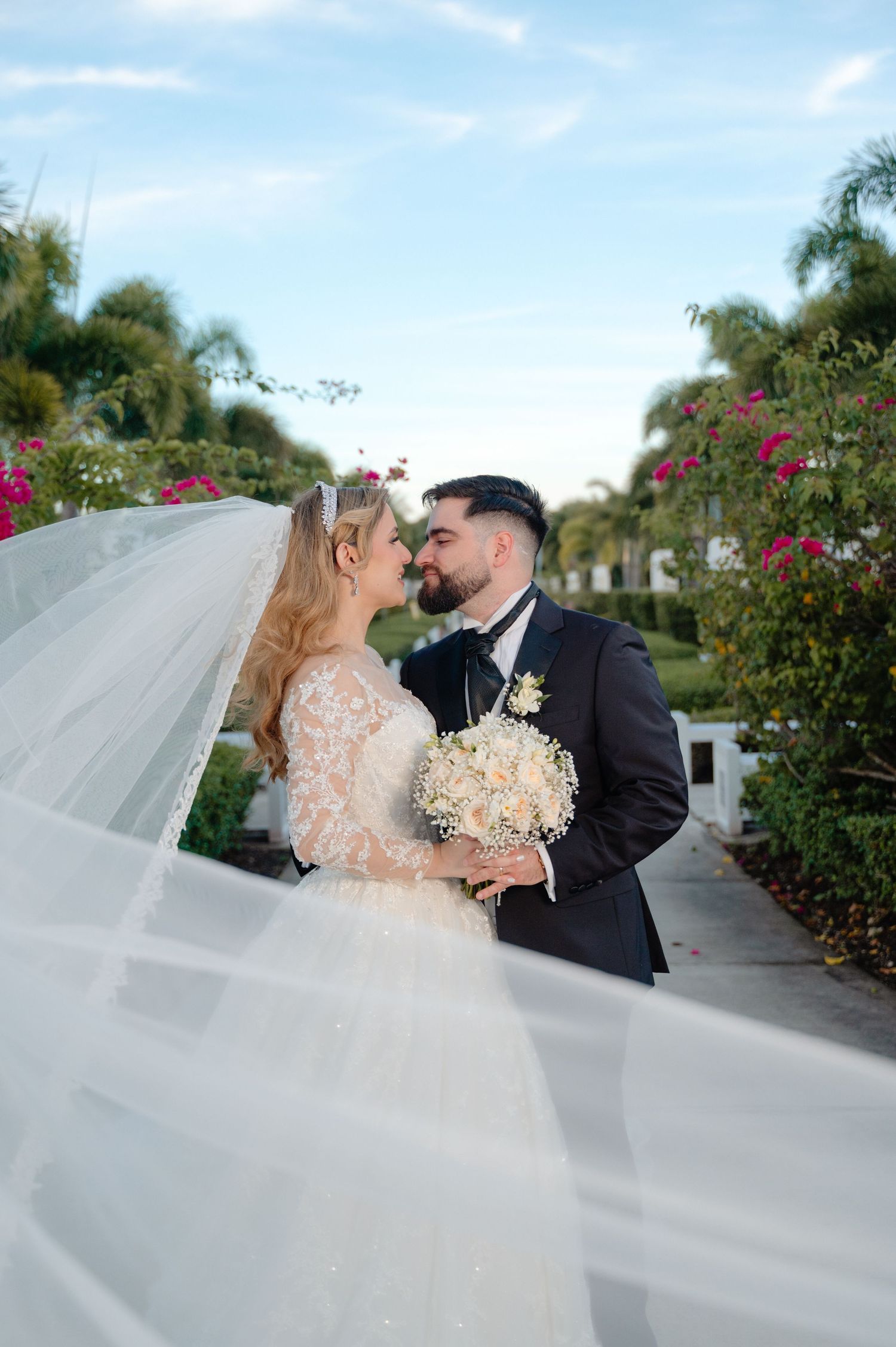 A romantic wedding moment captured on a palm-lined path with flowing veil and ivory bouquet.