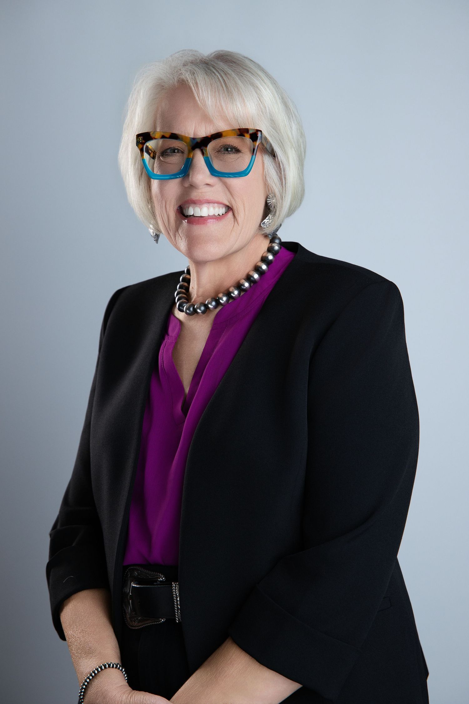 Professional woman with white hair, colorful glasses, black blazer, and purple blouse smiling confidently.