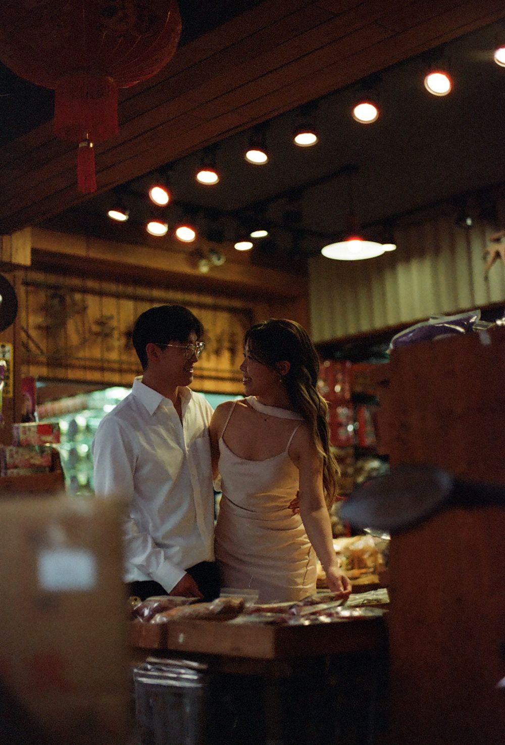 A couple shares a romantic moment in a warmly lit Asian market with red lanterns and glowing ceiling lights.