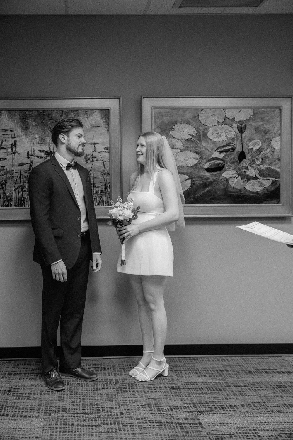 Black and white photo of a bride in short white dress holding bouquet and groom in tuxedo at courthouse wedding.