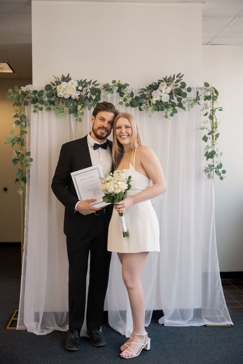 Smiling newlywed couple posing with marriage certificate and white bouquet in front of floral wedding backdrop.