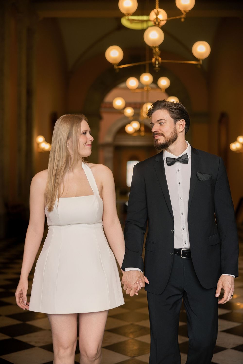 Bride in white mini dress and groom in black tuxedo hold hands walking in elegant chandelier-lit hall.