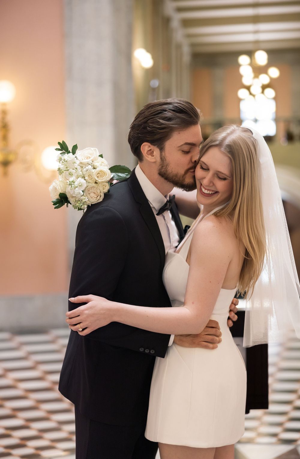 Bride and groom share a romantic kiss in elegant wedding attire, bride holding white roses bouquet.