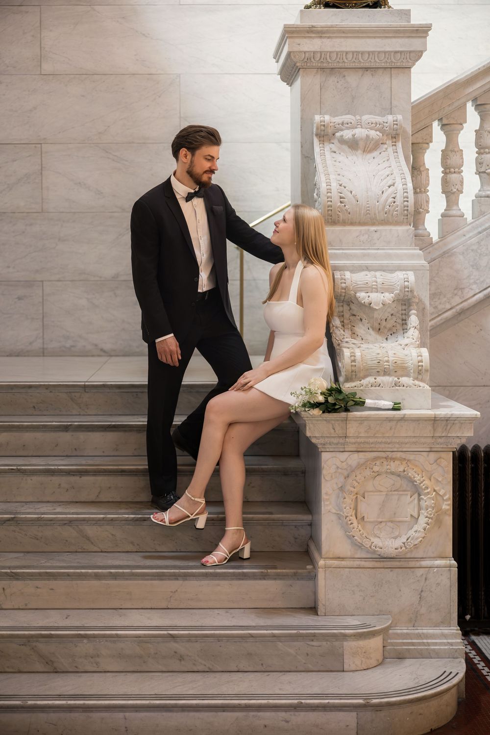 Elegant couple in formal attire on marble stairs, groom in tuxedo standing beside seated bride in white mini dress.