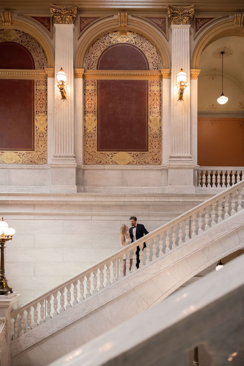 Couple in wedding attire sharing a romantic moment on grand marble staircase in elegant historic building.